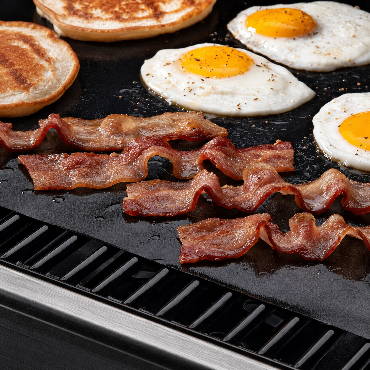 Eggs, bacon and toasted bread cooking on a black non-stick BBQ grill mat over an outdoor grill